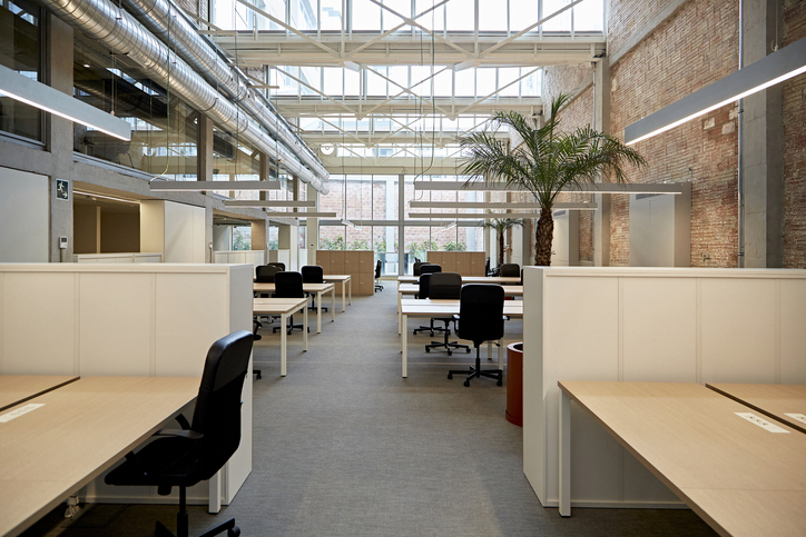 Wide angle view of unoccupied desks and chairs on ground floor of renovated building with exposed brick wall, high ceiling, and visible second floor windows.